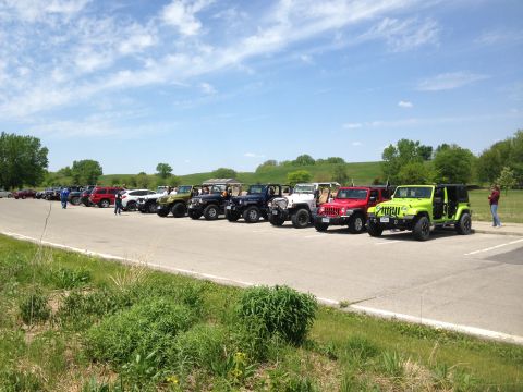 1979 Ford Bronco - A group of Jeeps are parked in a lot under a partly cloudy sky, with people milling about.