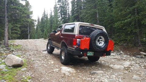 1996 Ford Bronco - A maroon Ford Bronco with a tan top is seen from the rear driving on a rocky, wooded trail.