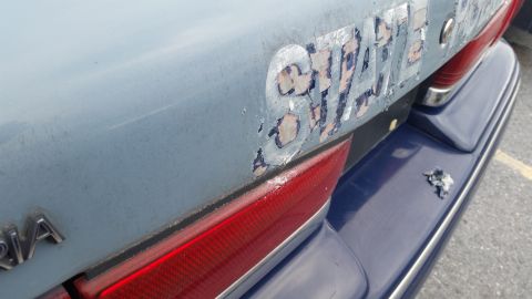 1991 Ford LTD Crown Victoria - The rear of a light blue car shows significant wear with peeling stickers and a damaged dark blue bumper.