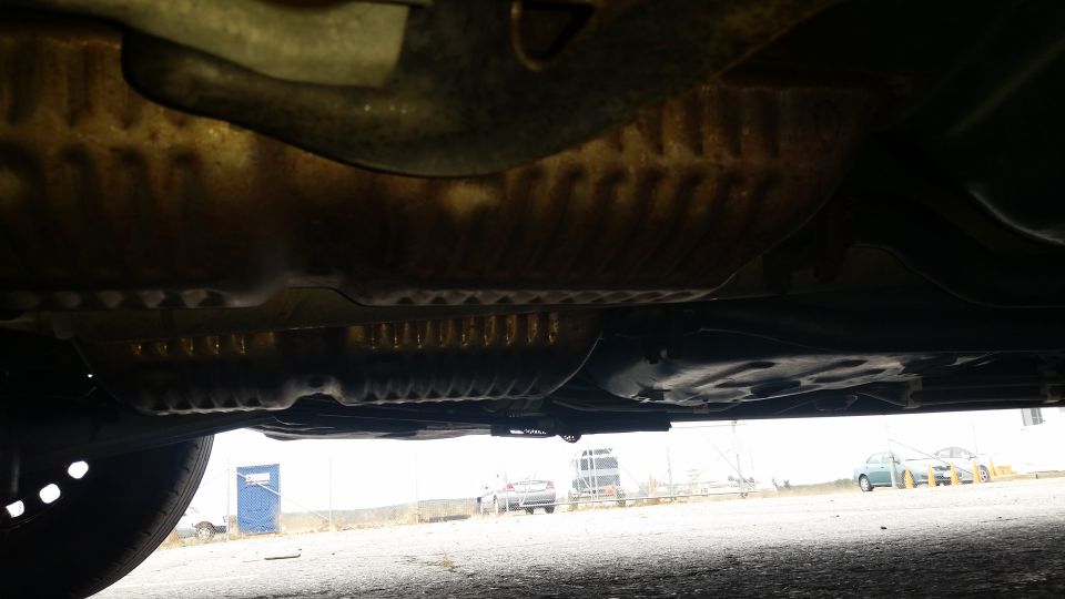 Underside of a car, showing exhaust and frame components. Other cars visible in background