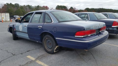 1991 Ford LTD Crown Victoria - A row of police cars, one with a flat tire, parked in a lot under an overcast sky.