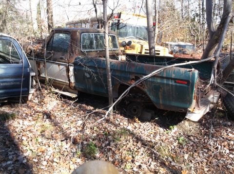 1974 Dodge Challenger - Multiple old trucks sit abandoned in a wooded area, overgrown with vegetation.