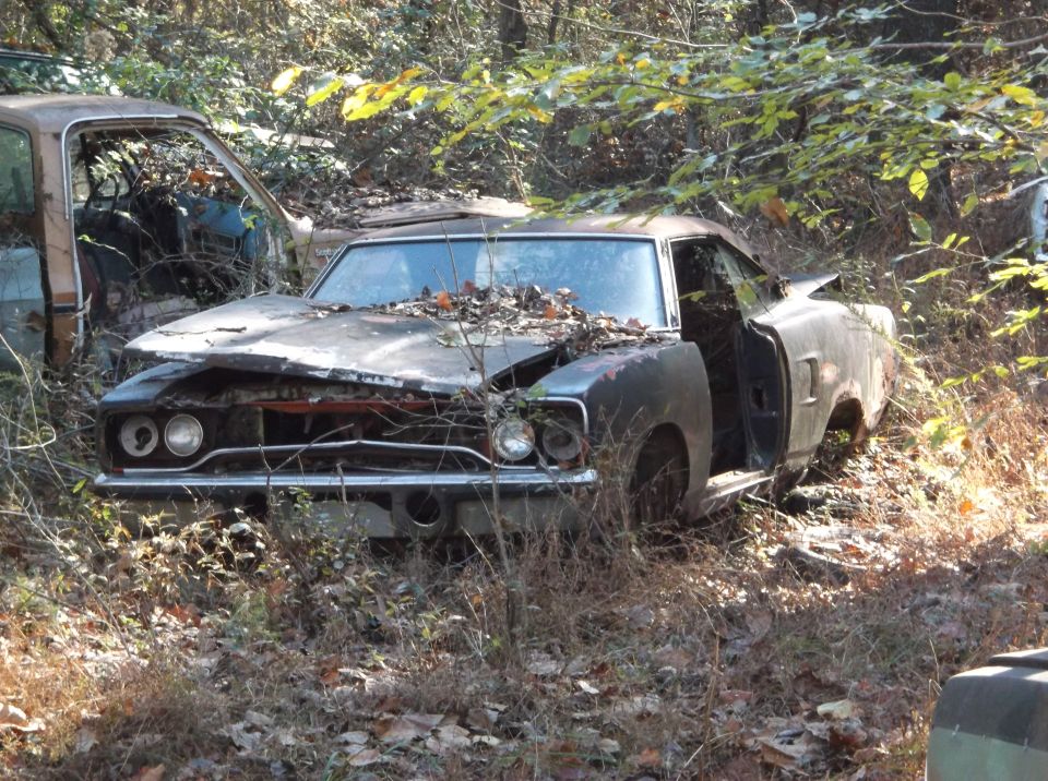 Old muscle car sitting in the woods, leaves on hood, driver door open. Another car is partially visible on the left