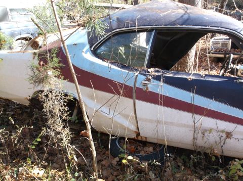 1974 Dodge Challenger - This abandoned vehicle, possibly a Dodge Challenger, sits in a wooded area with visible rust and overgrowth.