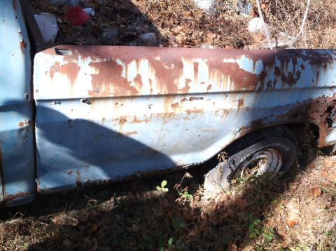 1974 Dodge Challenger - A heavily rusted blue pickup truck fender sits in overgrown brush, showing significant wear and tear.