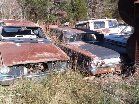 1974 Dodge Challenger - Several old, rusted vehicles are parked in a grassy junkyard under a clear sky.