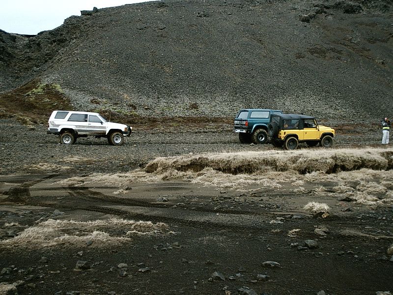 White, blue and yellow off-road SUVs parked on volcanic gravel below a scree slope