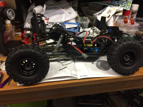 1989 Ford Bronco - RC rock crawler chassis with large tires sits on a workbench next to an open instruction manual.