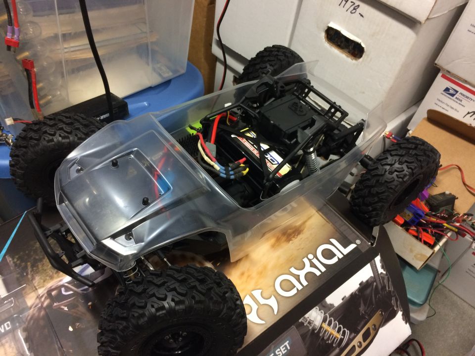 RC crawler chassis with clear body, battery, and wiring visible. Axial box underneath