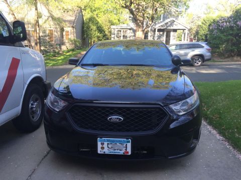 2013 Ford Taurus - A black 2013 Ford Taurus police car is parked in front of a house, with a white van to its left and a silver SUV in the background.