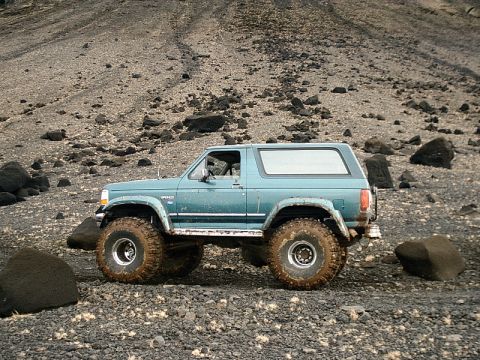 1996 Ford Bronco - This teal SUV, identified by its Bronco badging, is equipped with large tires for off-road use on a rocky landscape.