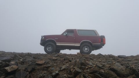 1996 Ford Bronco - A red 1996 Ford Bronco is parked on a rocky mountain peak, partially obscured by fog.