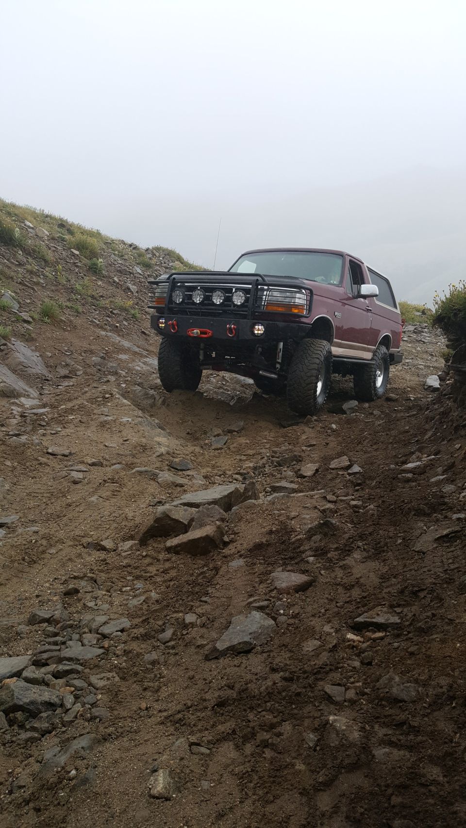 Maroon Ford Bronco climbing a rocky, steep dirt trail with a brush guard and auxiliary lights