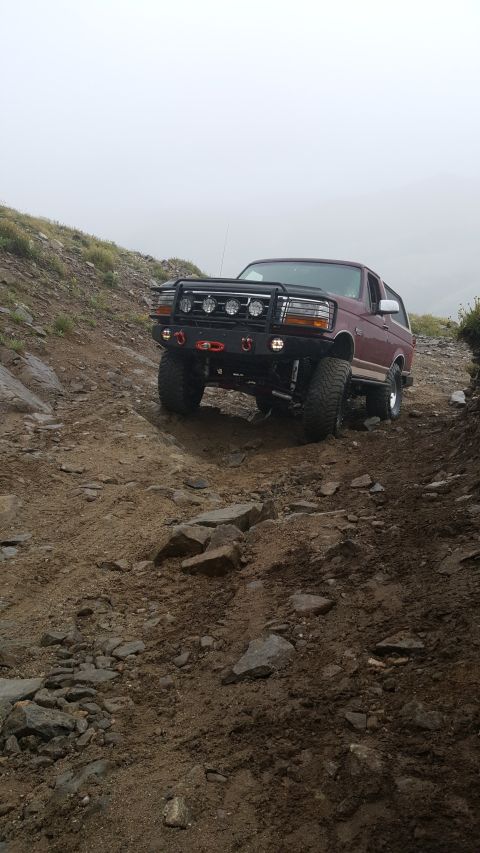 1996 Ford Bronco - A maroon Ford Bronco navigates a challenging, rocky off-road path under a hazy sky.