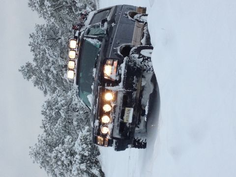 1996 Ford Bronco - A dark SUV is partially buried in snow, with its off-road lights illuminated against the wintery backdrop.