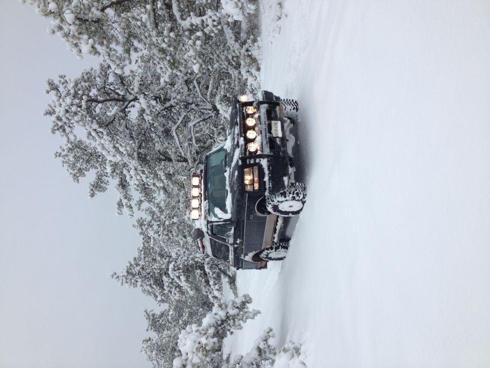 Dark SUV with multiple light bars on roof and bumper, driving in snow. Tires have snow chains