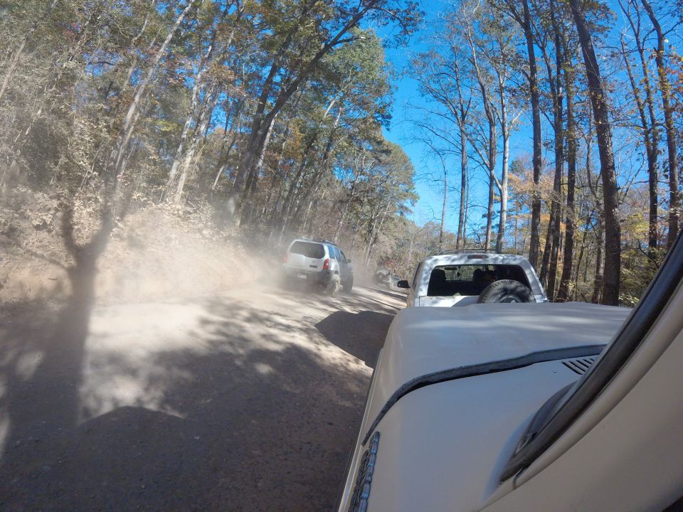 Two off-road vehicles driving on a dusty dirt road through trees. One vehicle is in the foreground, partially visible