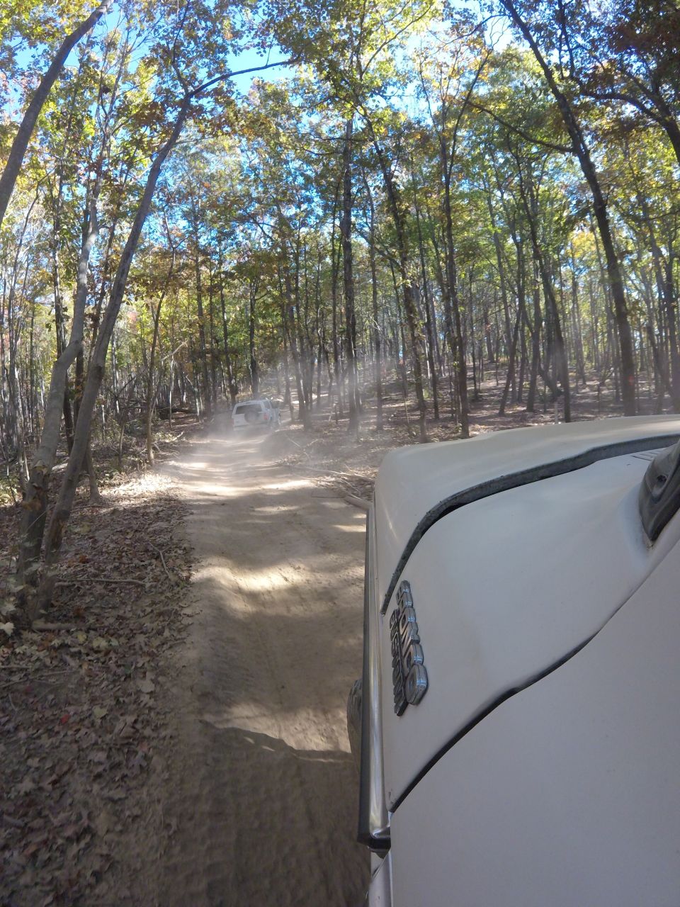 White SUV driving on a dusty forest road, another white SUV in the distance