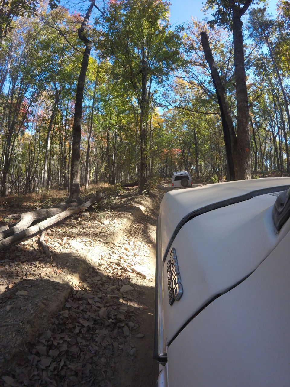White 1979 Ford Bronco driving on a dirt trail in a forest, another SUV follows behind