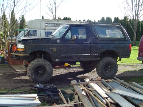 1993 Ford Bronco - This lifted 1993 Ford Bronco features oversized tires and a dark blue finish, parked outdoors with some debris around.