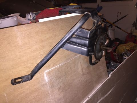 1967 Ford Custom Wagon - A black metal bracket with an oval mounting hole is angled across a piece of plywood. The bracket appears to be part of a larger assembly,…