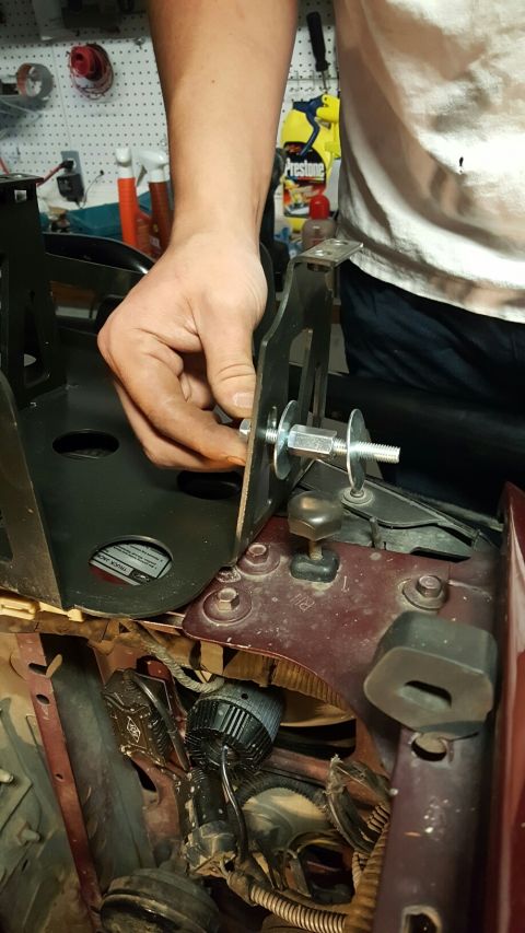 1996 Ford Bronco - A person's hand is shown installing a black metal bracket onto a vehicle's engine bay using a bolt, washer, and nut. The bracket appears to…