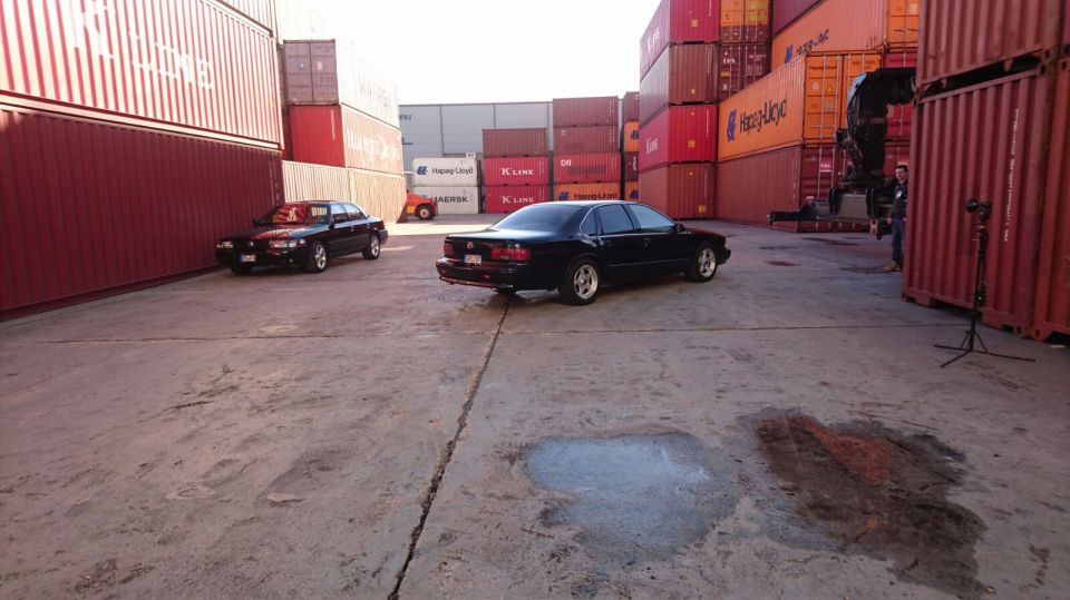 Two black sedans parked in a shipping container yard with a camera on a tripod.