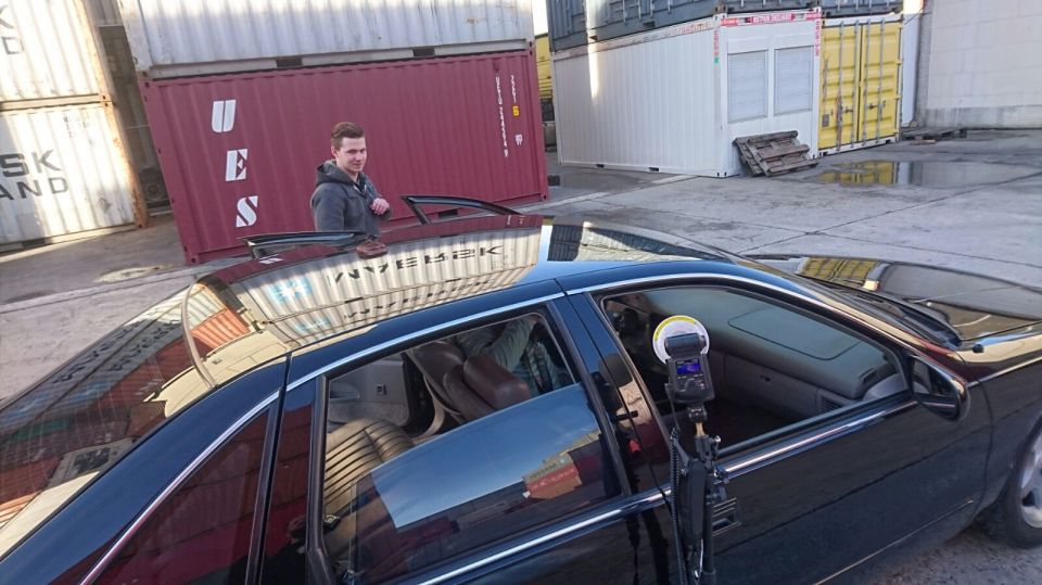 Black sedan with shipping containers in background, man leaning on door, camera flash inside.