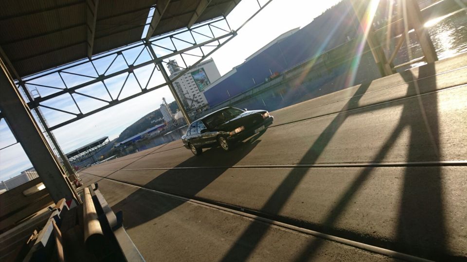 Black sedan parked on tracks under industrial structure with sun glare.