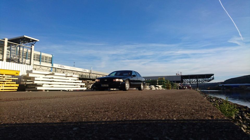 Black sedan parked outdoors in front of industrial buildings and stacked materials.