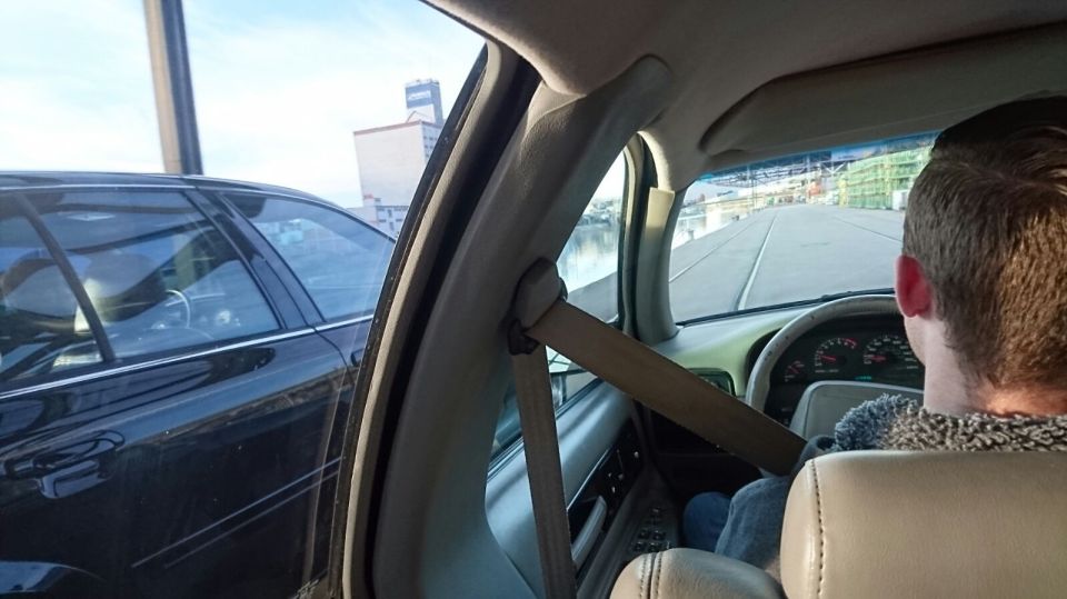 Interior of a dark sedan, driver with seatbelt on, view of road and industrial area.