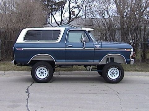 1995 Ford Bronco - Driver-side profile of a blue late-1970s full-size Ford Bronco with a white removable hardtop, parked on a residential street. It sits lift…