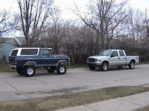 1995 Ford Bronco - Two Ford vehicles, a Bronco and a pickup truck, are parked on a street with bare trees in the background.