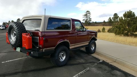 1996 Ford Bronco - The rear driver's side of a maroon and tan Ford Bronco is shown parked on asphalt next to a concrete curb. A spare tire and two red gas can…