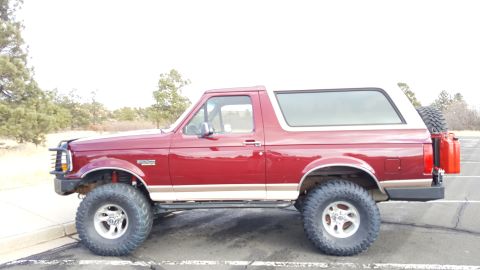 1996 Ford Bronco - A red 1996 Ford Bronco is parked on an asphalt surface with a white sky and trees in the background. The SUV has large off-road tires, a sp…