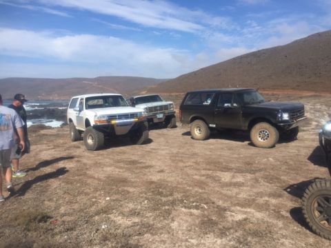 1986 Ford Bronco - Two lifted Ford Broncos are parked on a dirt lot near the coast, with a third vehicle partially visible on the right. The white Bronco is i…