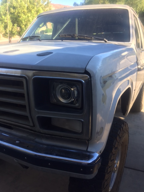 1986 Ford Bronco - The front corner of a white Ford Bronco is shown, with a chrome bumper and a modified headlight assembly. The vehicle appears to be undergo…