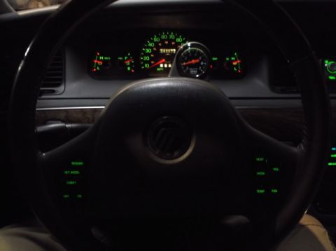 1999 Mercury Grand Marquis - The interior of a car shows a steering wheel with cruise control buttons and a dashboard with illuminated gauges. A tachometer is mounted o…