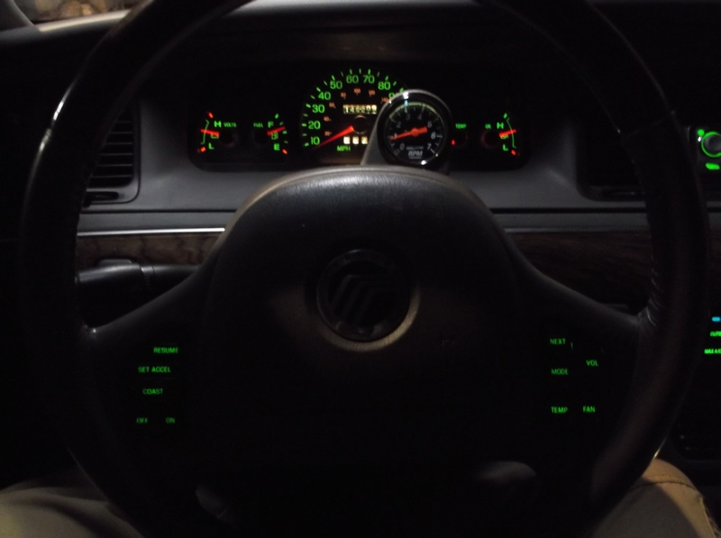 Interior view of a car dashboard and steering wheel with illuminated gauges and a tachometer.
