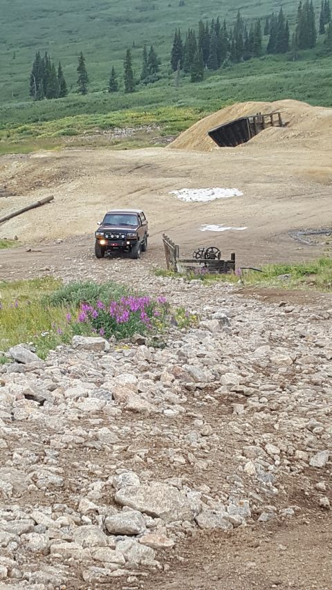 1996 Ford Bronco - A dark red 1996 Ford Bronco is parked on a rocky dirt road in a mountainous area with pine trees in the background. The Bronco has aftermar…