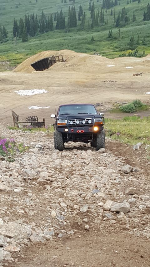 1996 Ford Bronco - A dark colored Ford Bronco drives up a rocky, mountainous dirt road. The truck has a custom front bumper with auxiliary lights and a winch.