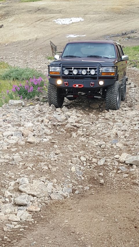 1996 Ford Bronco - A dark red 1996 Ford Bronco is parked on a rocky, off-road trail with purple wildflowers to its left. The Bronco features a black aftermark…