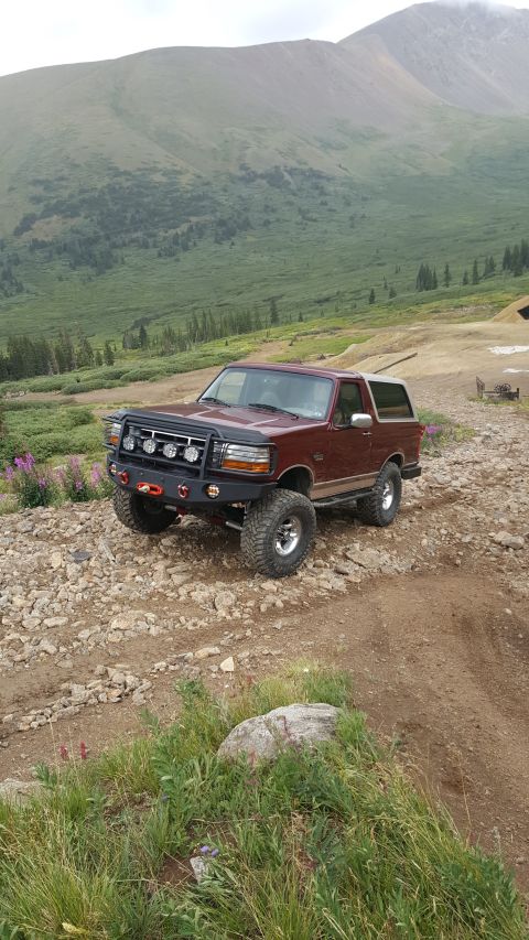 1996 Ford Bronco - A maroon 1990s Ford Bronco with aftermarket modifications is parked on a rocky, mountainous trail. The vehicle features a custom bumper wit…