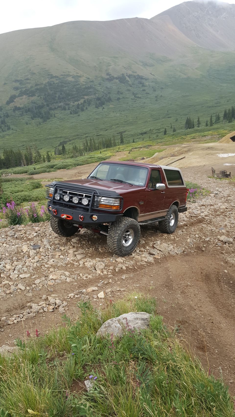 Maroon 1990s Ford Bronco with custom bumper and winch on a rocky mountain trail.