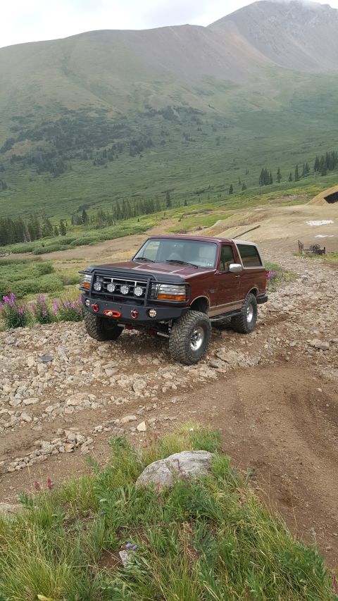 1996 Ford Bronco - A maroon 1996 Ford Bronco with a custom front bumper, winch, and auxiliary lights is parked on a rocky, mountainous trail.