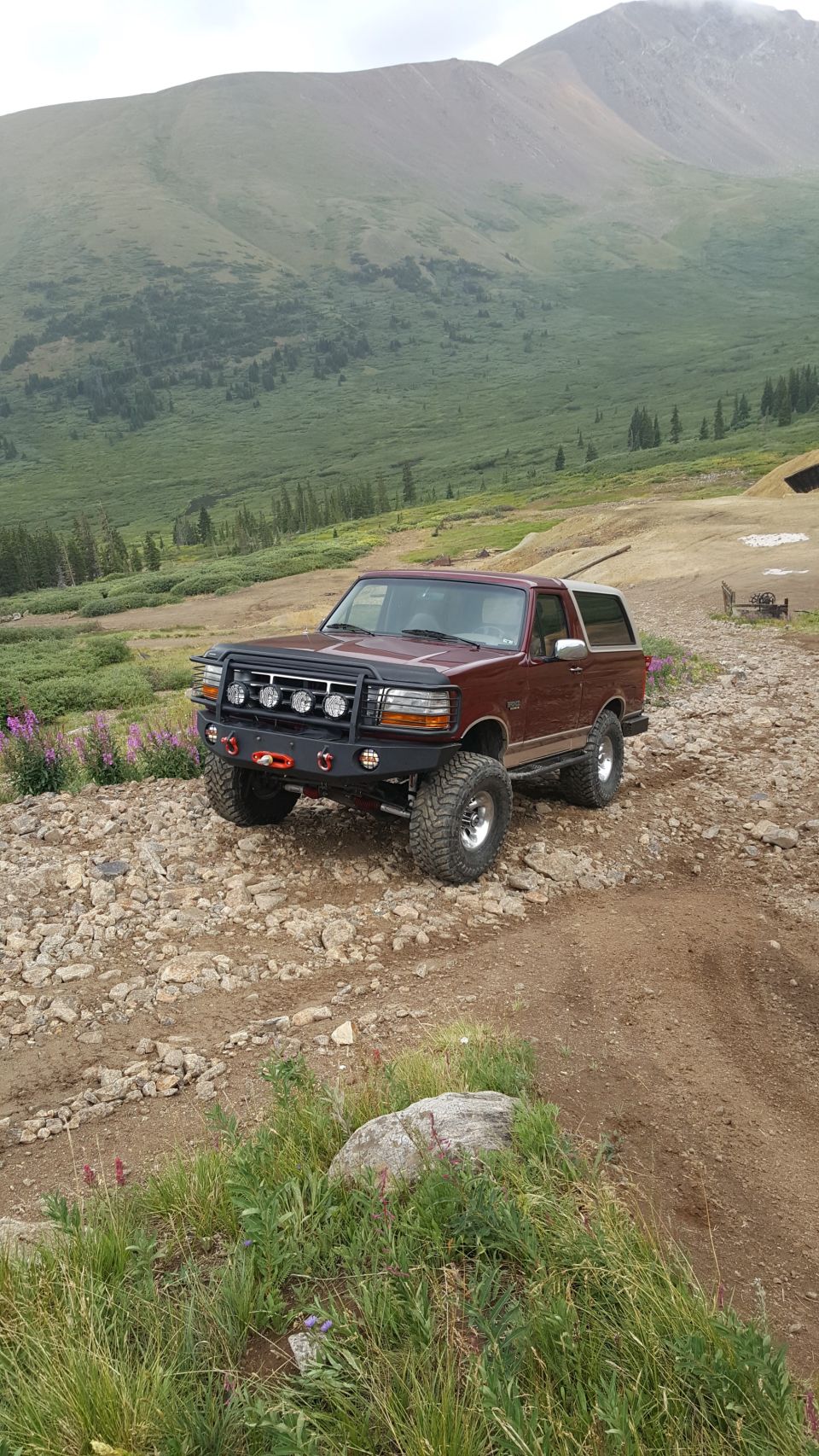 Maroon 1996 Ford Bronco with custom bumper and lights on a rocky mountain trail.