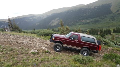 1996 Ford Bronco - A maroon 1996 Ford Bronco is shown in a three-quarter front view, driving on a rocky, off-road trail in a mountainous, forested area. A red…