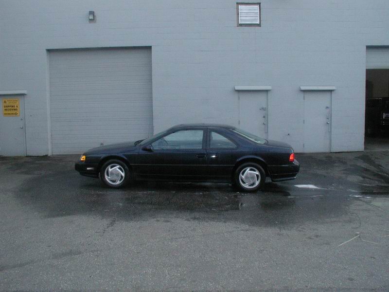 Dark blue 1990s Ford Thunderbird coupe in profile on wet pavement by a white warehouse