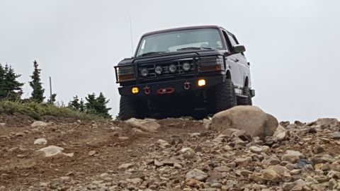 1996 Ford Bronco - A dark-colored Ford Bronco is shown from a low front-three-quarter angle, climbing a rocky, off-road trail. The vehicle features an afterma…