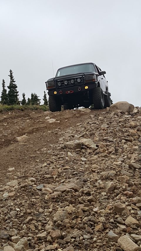 1996 Ford Bronco - A dark-colored Ford Bronco is parked on a rocky, uphill trail with trees in the background and a cloudy sky overhead. The vehicle has a cus…